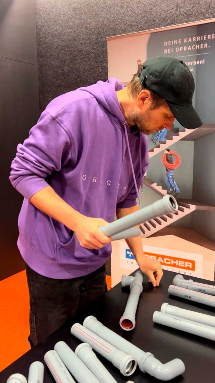 Person in violettem Hoodie und schwarzer Kappe begutachtet graue PVC-Rohre und Formstücke auf einem Tisch an einem Messestand.